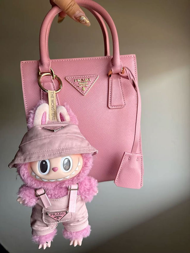  Labubu em look rosa pelúcia com chapéu e jardineira, pendurado em uma bolsa rosa da Prada.