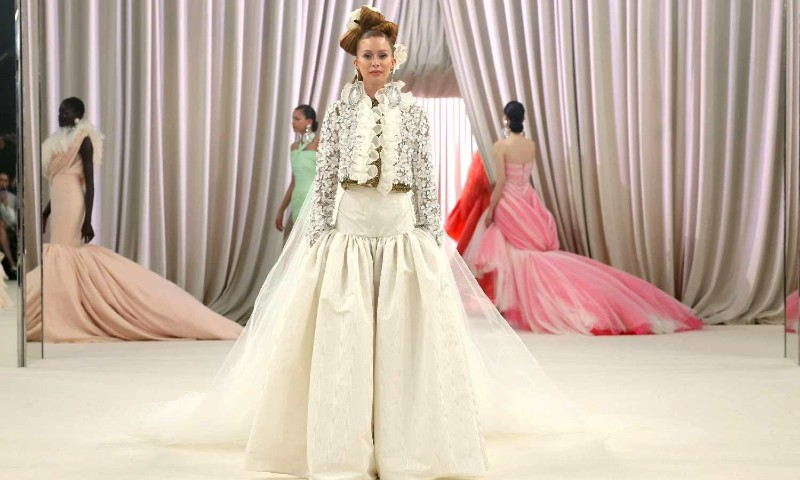 Modelo desfilando em uma passarela com um vestido branco volumoso de alta-costura. A peça tem uma saia ampla e estruturada, combinada com uma jaqueta curta bordada com detalhes florais e babados. O cabelo está preso em um penteado elaborado com uma flor branca como acessório. Ao fundo, duas outras modelos vestem vestidos sofisticados em tons de rosa e salmão, posando diante de cortinas drapeadas em um cenário elegante.