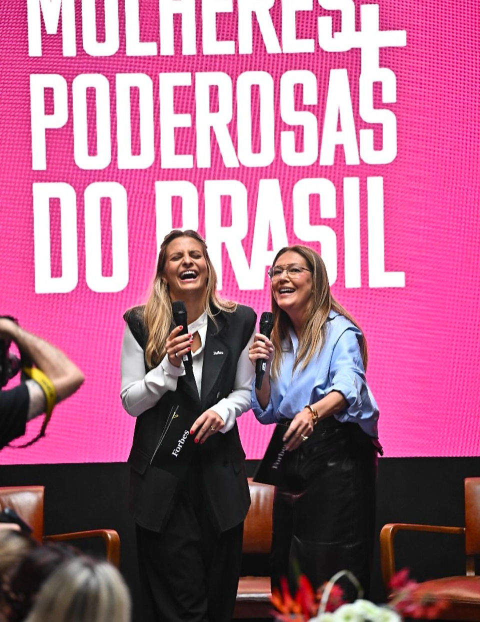 Duas mulheres sorrindo com microfones no palco, em frente a um painel rosa com os dizeres “Poderosas do Brasil”.