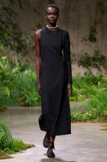 Uma modelo desfila pela passarela no desfile da Gucci em Londres, usando um vestido preto minimalista e elegante, de comprimento midi e com uma manga longa assimétrica. O look é complementado com um colar de pérolas e sapatos pretos com detalhes em laço. O cenário da passarela é coberto por vegetação verde exuberante, criando um contraste sofisticado com o visual monocromático da modelo.