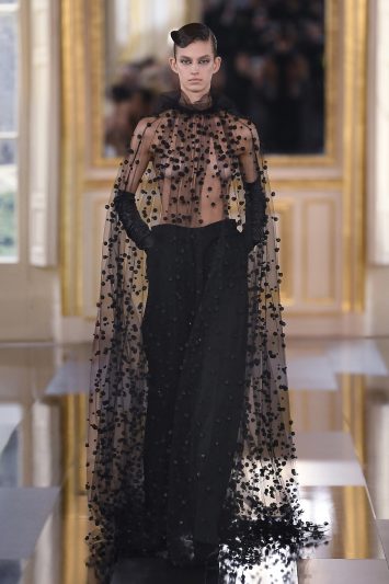 Uma modelo desfila com um look da Valentino composto por uma blusa transparente de tule adornada com detalhes bordados em pequenos pompons pretos e uma capa longa igualmente decorada, criando um efeito etéreo. A peça é combinada com calças pretas de alfaiataria de cintura alta e luvas longas. A modelo exibe uma expressão séria e uma postura elegante enquanto caminha pela passarela espelhada, refletindo o cenário luxuoso com molduras douradas ao fundo durante a Paris Fashion Week