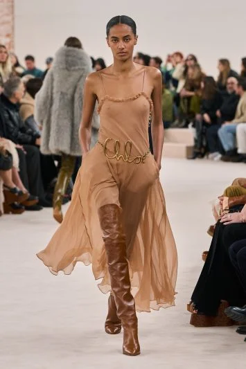 Modelo desfilando com um vestido fluido em tom nude, com detalhe de cinto dourado formando a palavra 'Chloé', combinado com botas de cano alto marrons, em uma passarela com plateia ao fundo