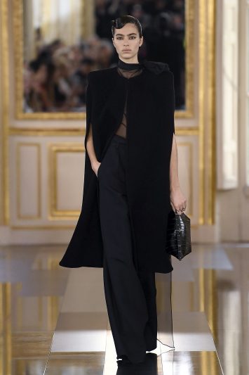 Uma modelo desfila com um look preto sofisticado composto por uma capa longa estruturada, calça de alfaiataria ampla e blusa de gola alta semi-transparente durante a Paris Fashion Week. A modelo carrega uma bolsa preta estruturada e mantém uma expressão séria enquanto caminha pela passarela espelhada, refletindo o ambiente clássico com molduras douradas ao fundo