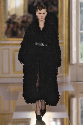 Uma modelo desfila vestindo um longo casaco preto com textura felpuda volumosa, marcado na cintura por um cinto preto de couro com fivela prateada. O look, que transmite uma silhueta estruturada e impactante, é complementado por sapatos pretos de salto. A modelo mantém uma expressão séria e confiante enquanto caminha pela passarela espelhada, com o cenário luxuoso de molduras douradas ao fundo durante a Paris Fashion Week