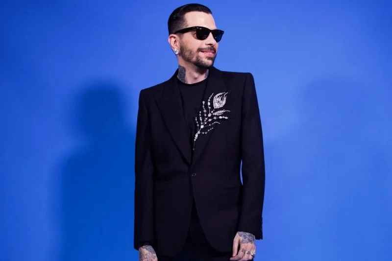 O designer Eduardo Claires, responsável pelos acessórios da coleção, posa com um look preto sofisticado, adornado com um bordado prateado em forma de pena, trazendo um toque de elegância discreta.