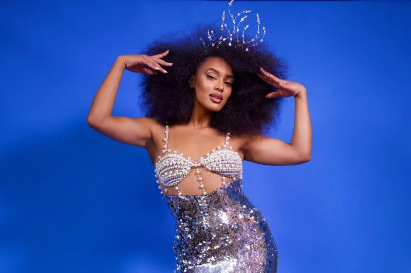 Raissa Santana exala empoderamento com um vestido deslumbrante de paetês prateados e pérolas, coroado por um acessório de cabeça elegante que reforça o brilho e glamour do look.