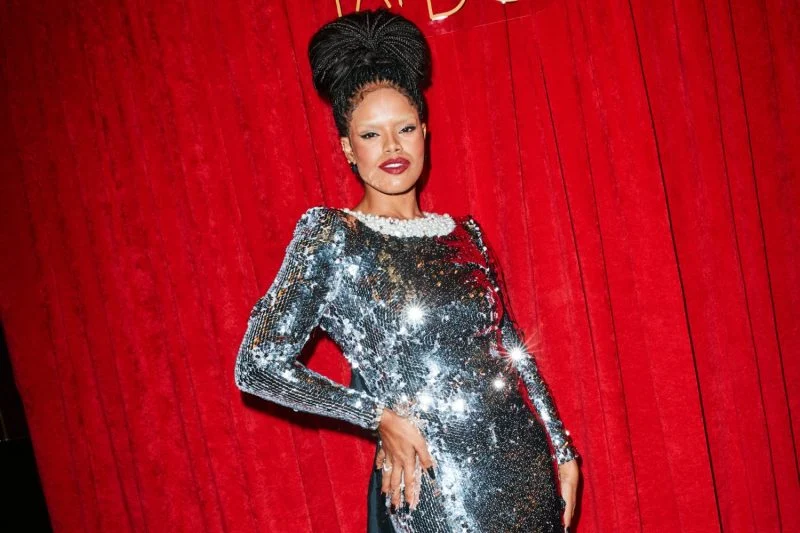 Josy Ramos brilha em um vestido justo de paetês prateados com ombros estruturados e gola adornada com pérolas, em frente a um fundo vermelho vibrante que destaca ainda mais seu glamour.