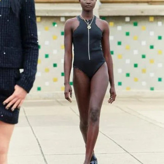 Modelo desfilando em um maiô preto com design minimalista, alças finas e um zíper frontal. O look é complementado por um colar com pingente dourado e sapatilhas pretas. Ao fundo, o cenário apresenta um prédio moderno com azulejos coloridos em tons de verde e amarelo. Outra modelo aparece parcialmente à esquerda, vestindo um conjunto preto texturizado