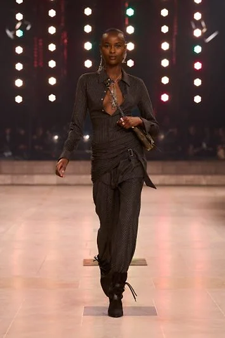 Modelo em um conjunto risca-de-giz marrom escuro, com blusa de decote profundo e calças ajustadas, combinado com botas pretas.