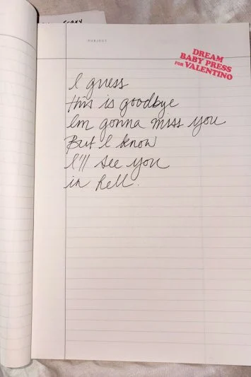Um caderno de capa dura aberto sobre um tecido branco, com um bilhete escrito à mão que diz: "I guess this is goodbye, I’m gonna miss you, but I know I’ll see you in hell." A marca Dream Baby Press for Valentino está impressa no canto superior direito.