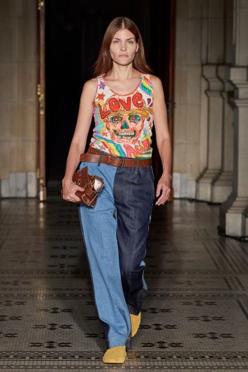 Modelo desfila com calças jeans de pernas de cores diferentes, e regata colorida com estampa de caveira.