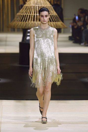 Modelo desfilando na passarela da Dior com vestido curto dourado coberto de franjas metálicas brilhantes, combinando com sandálias delicadas e cenário decorado com estruturas de junco iluminadas