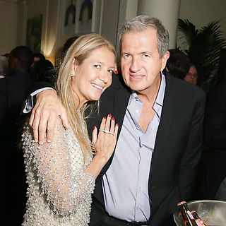  Um casal posa em um evento. A mulher, loira e vestindo um vestido de mangas longas com detalhes brilhantes, apoia-se no homem, que veste um terno preto e camisa azul. O ambiente é elegante, com uma bandeja de garrafas ao fundo.