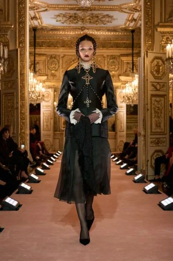 Modelo em um conjunto de alta-costura de inspiração vitoriana, combinando blazer de seda preta com bordados dourados, punhos rendados e uma saia de tule translúcido, evocando um estilo gótico elegante.