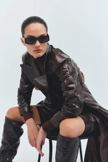 Modelo com expressão imponente posa sentada em um banco, vestindo um casaco texturizado de couro marrom com brilho, botas de cano alto combinando e óculos de sol escuros. O look sofisticado e futurista transmite uma estética poderosa, com styling minimalista e atitude forte. O fundo claro destaca o contraste do visual, reforçando a presença marcante da peça.