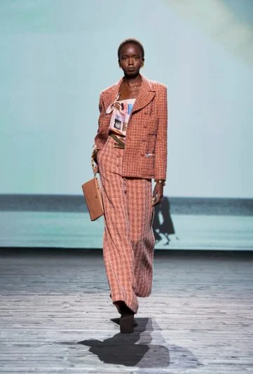 Modelo no desfile Chanel FW 2024/25, vestindo um conjunto de tweed rosa com blazer oversized e calça ampla, complementado por uma clutch bege e camiseta estampada, desfilando em uma passarela com cenário de praia serena