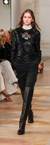 Suéter preto bordado com flores e paetês, usado com gola alta rendada, calça de alfaiataria e botas de couro cano alto em um look que mistura romantismo e sobriedade na passarela de Ralph Lauren.