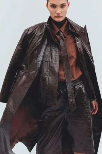 Modelo com expressão intensa e olhar fixo veste um conjunto de couro marrom texturizado com brilho, composto por um trench coat esvoaçante, bermuda ampla e camisa social combinando. A gravata fina adiciona um toque de sofisticação ao look poderoso e estruturado, evocando uma estética moderna e arrojada.