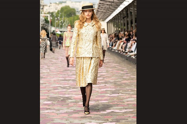 Modelo desfilando em uma passarela ao ar livre, trajando um conjunto dourado com textura rica e detalhes clássicos, complementado por um chapéu elegante de abas largas. O look exala sofisticação e charme vintage, enquanto o público ao fundo aprecia o desfile.