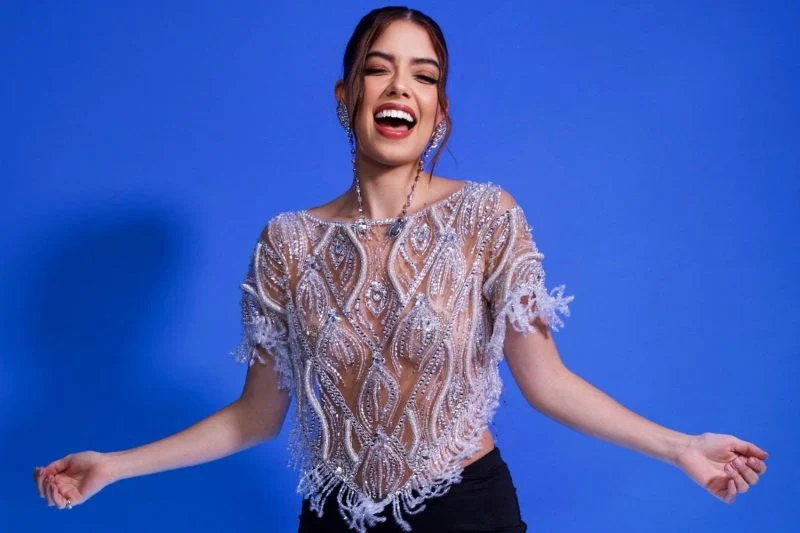 Vitória Fiore aparece radiante em um fundo azul vibrante, vestindo uma blusa transparente bordada com pedrarias brilhantes, combinada com saia preta. Seu sorriso ilumina a cena.