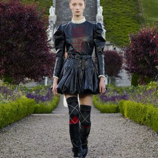 A imagem apresenta uma modelo desfilando em um jardim formal, vestindo um look composto por um vestido preto de couro com mangas bufantes e saia curta volumosa. A peça traz uma mensagem textual sutil no peito e é combinada com botas altas que exibem padrões de losangos em preto e vermelho, adicionando um toque gráfico ao visual. O cenário ao fundo, com escadarias e vegetação simétrica, equilibra a ousadia moderna do look com a elegância clássica do ambiente.