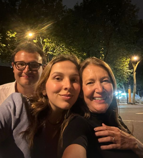 Três pessoas sorrindo para a câmera em uma foto noturna ao ar livre. À direita, Donata Meirelles veste preto e está com a mão sobre o ombro da pessoa ao centro. No meio, uma jovem de cabelos castanhos soltos veste blusa azul-clara, e à esquerda, um homem de óculos aparece parcialmente na imagem. O fundo mostra árvores e luzes de rua, criando um ambiente descontraído.
