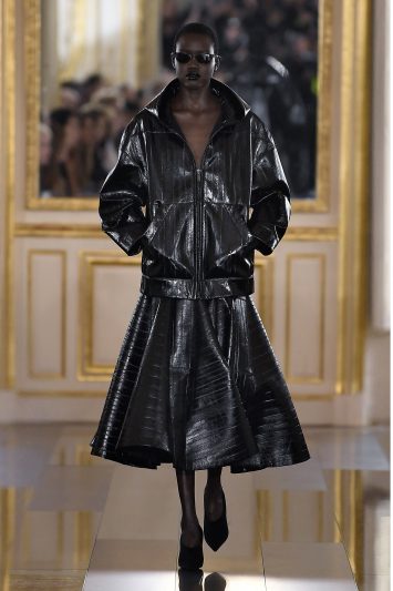 Uma modelo desfila com um conjunto preto brilhante de couro composto por uma jaqueta oversized com zíper e uma saia volumosa midi, criando uma silhueta dramática. Ela usa óculos escuros, sapatos de salto e mantém uma expressão forte e confiante enquanto caminha por uma passarela espelhada, com molduras douradas ao fundo durante a Paris Fashion Week