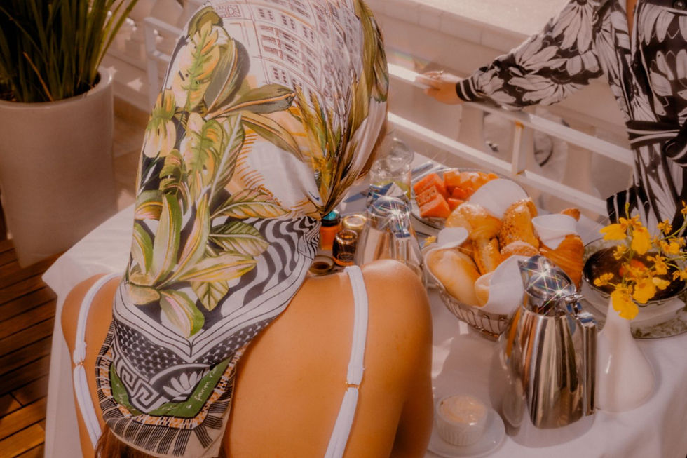 Mulher de costas usando um lenço estampado tropical na cabeça, sentado à mesa de café da manhã ao ar livre. A mesa está decorada com frutas frescas, pães e flores amarelas, em um ambiente iluminado pelo sol com detalhes elegantes ao fundo.