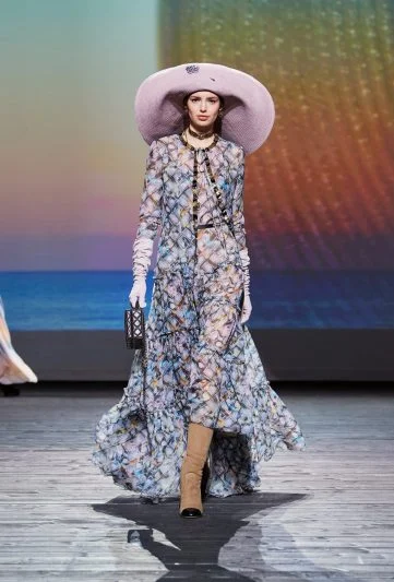 Modelo no desfile Chanel FW 2024/25, usando um vestido longo floral com transparência, chapéu rosa oversized, botas bege e luvas brancas, desfilando em uma passarela com cenário de pôr do sol à beira-mar