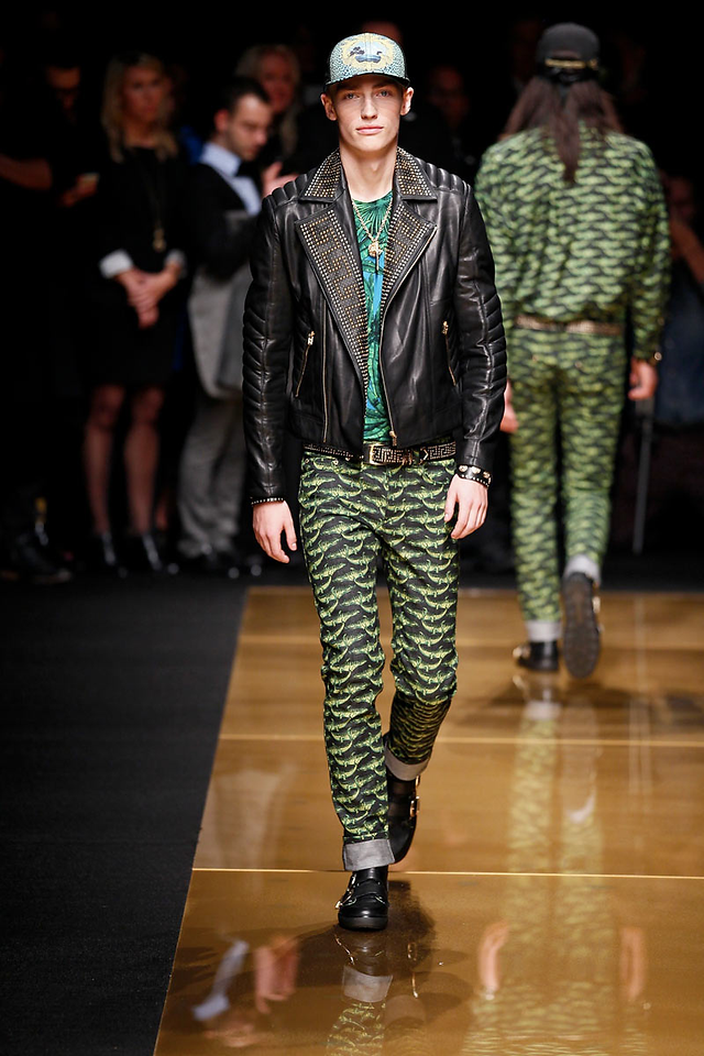 Modelo desfilando com conjunto verde estampado e jaqueta de couro preta em uma passarela com iluminação dourada.