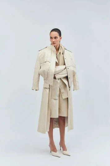 Modelo com expressão forte posa contra um fundo neutro vestindo um look monocromático em tons de bege. Ela usa um trench coat oversized estruturado, combinado com um conjunto de blazer e bermuda no mesmo tom, além de scarpins de bico fino. O styling minimalista e sofisticado traz uma abordagem moderna e elegante ao visual