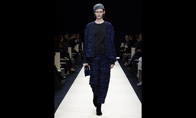  Modelo vestindo um conjunto de blazer e calça azul com textura bordada, boina azul combinando e segurando uma clutch preta, desfilando com um ar sério.
