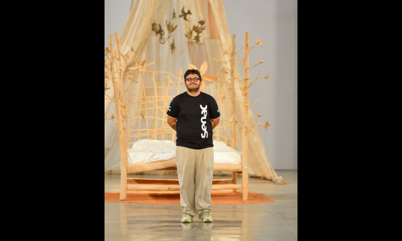 Antônio Castro agradece o público pela presença em seu desfile. Ele usa camiseta preta do Senac, calças beges, óculos redondos e tênis também beges.