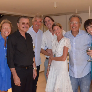 Donata Meirelles e um grupo de convidados posam sorridentes em um ambiente interno sofisticado. Todos vestem looks elegantes em tons claros e escuros, transmitindo uma atmosfera de celebração e requinte.