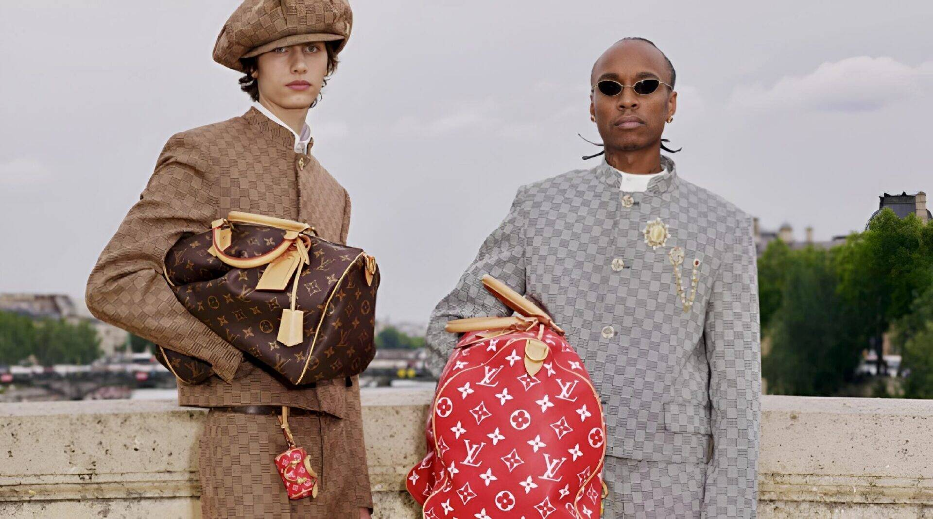 Dois modelos posando em um cenário parisiense, vestindo peças com o monograma LV em tons de marrom e cinza, segurando bolsas grandes com o icônico padrão da Louis Vuitton em vermelho e marrom.