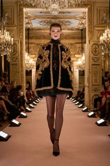 Modelo em uma capa curta preta e dourada, bordada com padrões barrocos, combinada com um mini vestido de renda e meia-calça, desfilando com confiança em um cenário suntuoso.