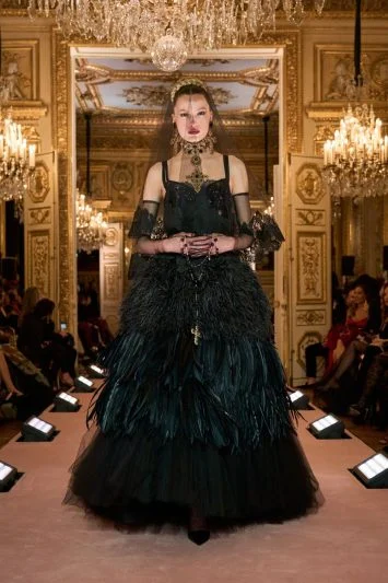 Modelo em um vestido preto volumoso, repleto de plumas e detalhes dourados, transmitindo um ar de sofisticação dramática enquanto caminha pela passarela luxuosa.