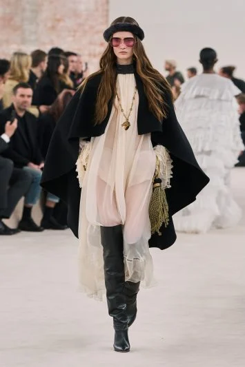 Modelo desfilando com um look composto por um vestido fluido branco, capa preta estilosa, botas de cano alto pretas, acessórios dourados e óculos de sol oversized, em uma passarela com plateia ao fundo