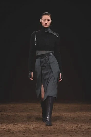 Modelo na passarela com um look preto composto por suéter de gola alta com detalhe de alça em couro e saia cinza com amarração lateral e fenda discreta, transmitindo elegância e força.