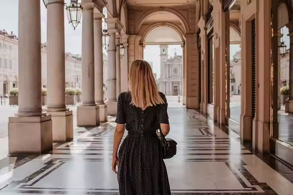 A woman wearing a black dress and carrying a purse walking outside on a tile floor away from the photographer