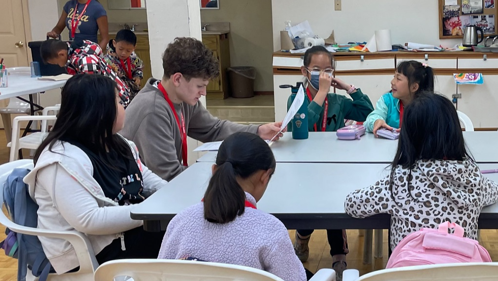 Children and a teacher sit at tables in a classroom, reading papers and talking; colorful bags and supplies in the background.