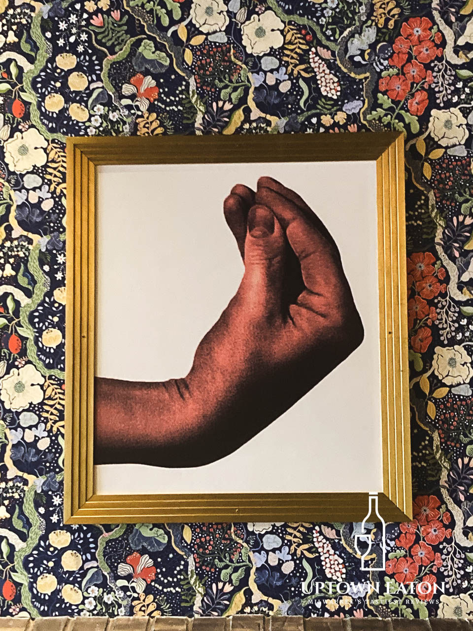 A framed artwork featuring a stylized image of a red-toned hand in the classic Italian gesture for “What do you want?” hangs against a dark floral-patterned wallpaper. The frame is gold with ridged detailing. 