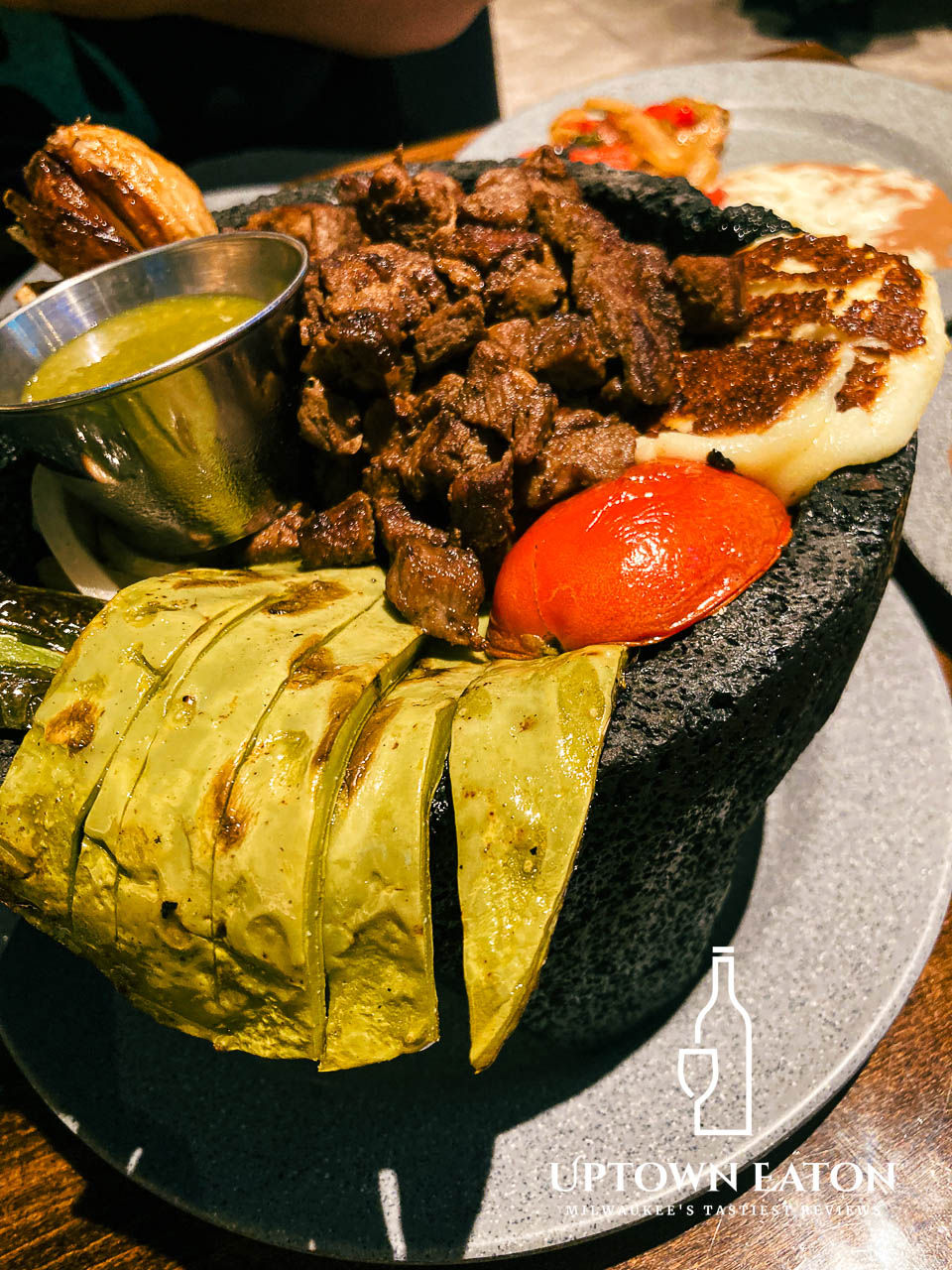 A plate with grilled meat, green sauce, toasted cheese, nopal, and a red tomato in a stone bowl.