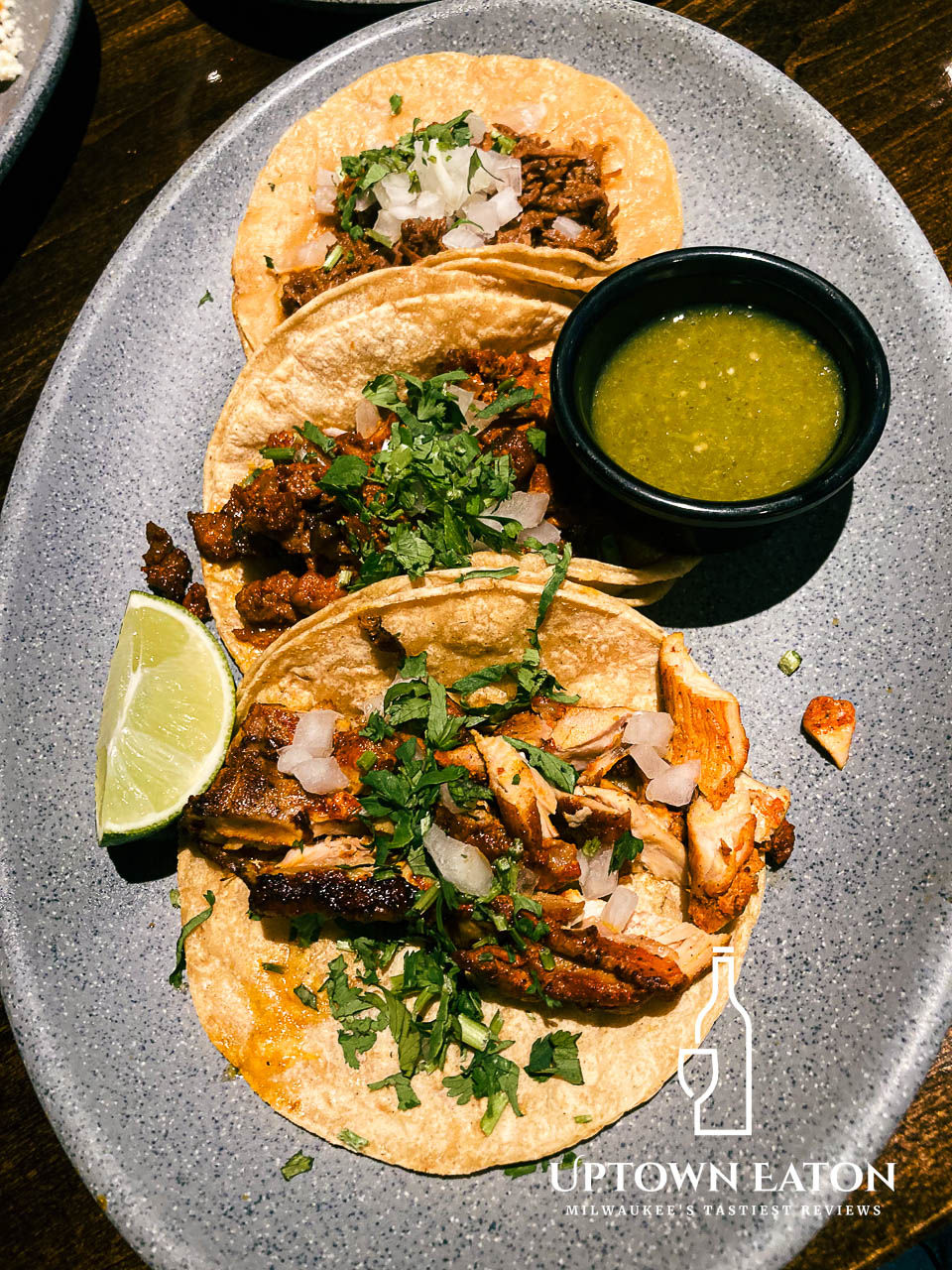 Three tacos with meat, cilantro, and onions on a plate with lime and green sauce. Dark table background.