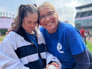 Two women smiling together, wearing blue shirts with logo, at baseball stadium Park Lawn