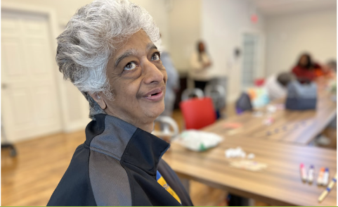 Smiling elderly woman looking at camera, seated at table, other people in background.