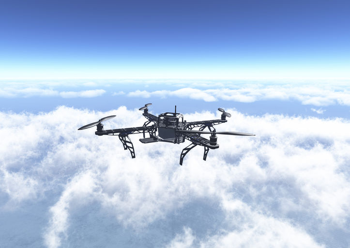 Black drone flying high above white clouds under a clear blue sky.