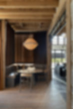 Rustic dining nook with a round table, plush bench, wooden chairs, and a layered chandelier. Dark curtains, wooden beams, white oak ceiling cladding, Natural Face Northern White Oak wide plank floors authentically crafted by Hardwood Design Company and outdoor view.