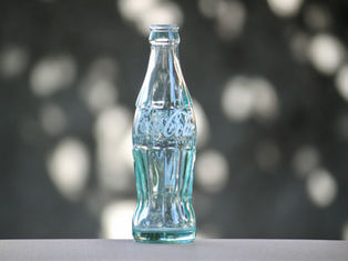 Clear Coca-Cola glass bottle on a neutral surface with a blurred, bokeh background. The text "Coca-Cola" is visible on the bottle.