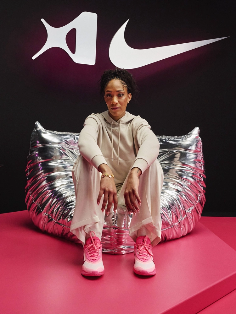 A'ja Wilson in beige outfit and pink sneakers sits on a silver chair. Black background with white Nike logos. Mood is relaxed and focused.